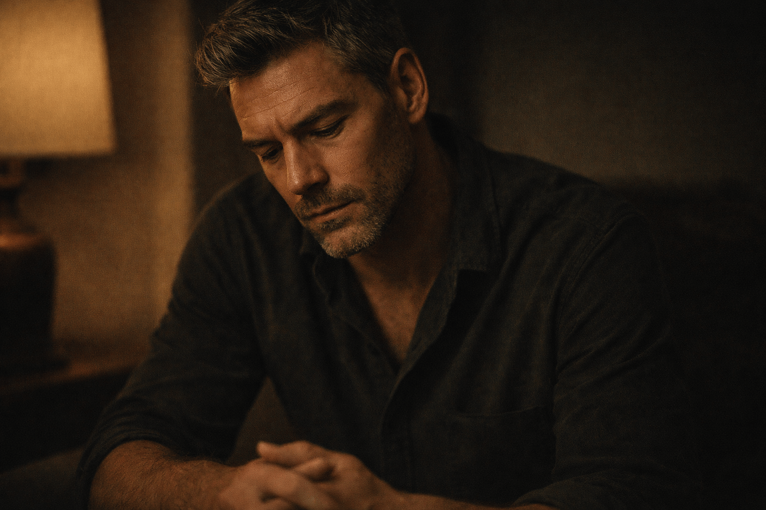 A man seated indoors in low, warm light, looking down thoughtfully with hands loosely clasped, conveying a quiet, reflective presence.